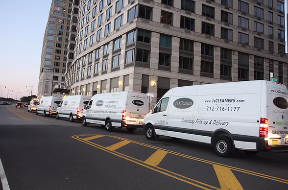 A row of white delivery vans with "J.C. Cleaners" logos are parked along the curb beside a tall office building in an urban area.