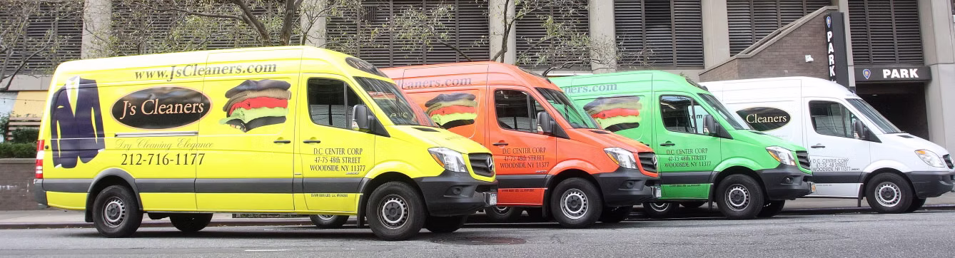 Four JS Cleaners vans in yellow, orange, green, and white are parked in a row along a city street in front of a building.