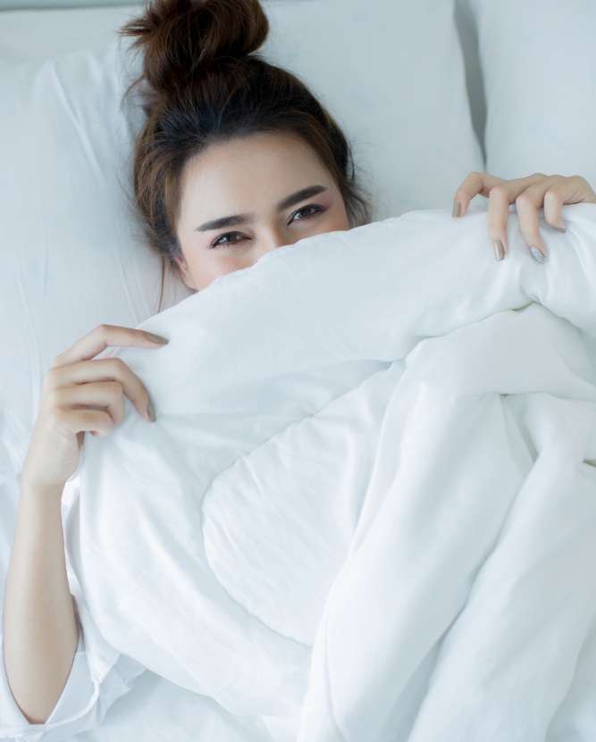 A woman lies in bed, mostly covered by a white blanket, with only her eyes and part of her face visible as she looks at the camera.