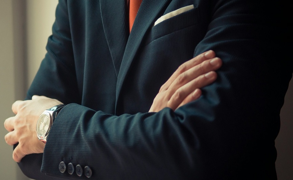 Person in a dark suit with an orange tie and pocket square stands with arms crossed, wearing a watch on their left wrist.