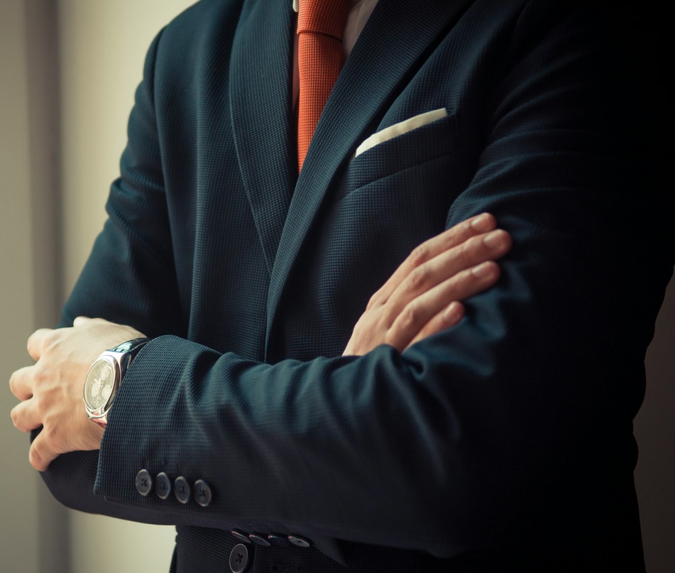 Man in a dark suit with an orange tie and white pocket square stands with arms crossed, wearing a wristwatch.