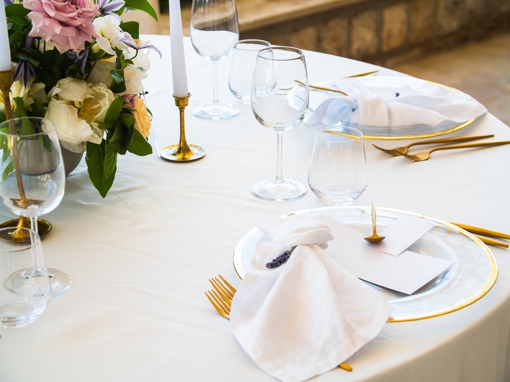 Elegant table setting with gold-rimmed plates, gold cutlery, wine glasses, white napkins, blank cards, and a floral centerpiece on a white tablecloth.