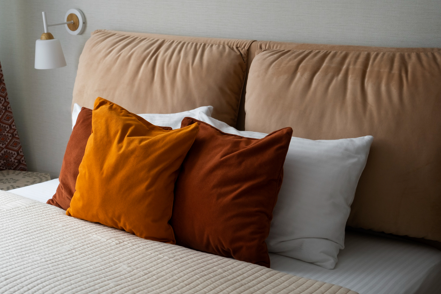 A neatly made bed with a beige headboard, two white pillows, and two brown and one orange decorative cushions in front.