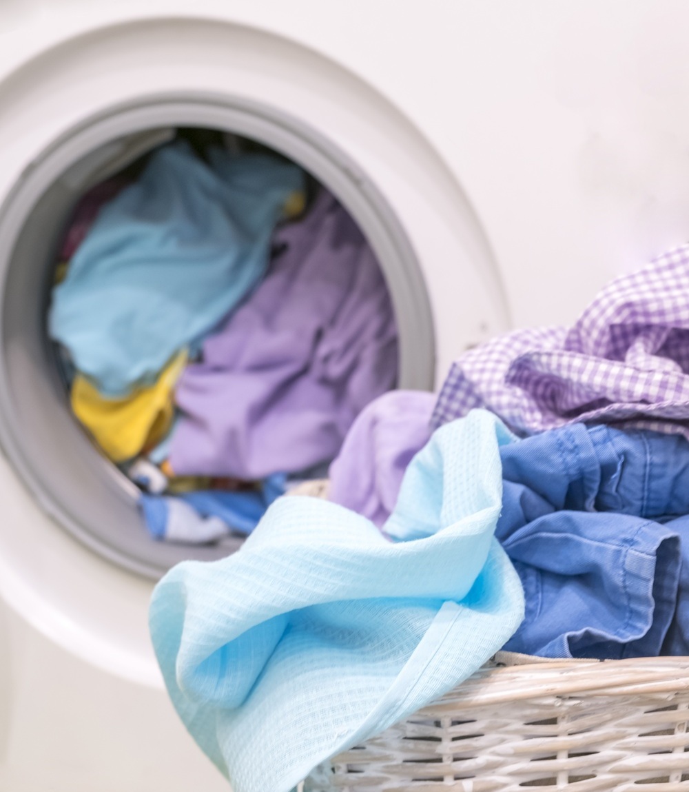 A front-loading washing machine filled with colorful clothes, with a laundry basket of clean clothes in the foreground.