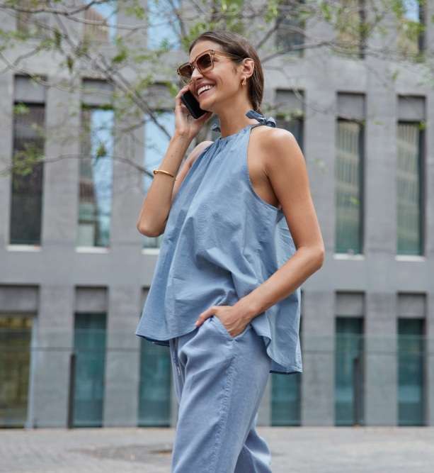 Woman wearing sunglasses and a loose blue top with matching pants stands outdoors, smiling and talking on a cellphone, with modern building windows in the background.