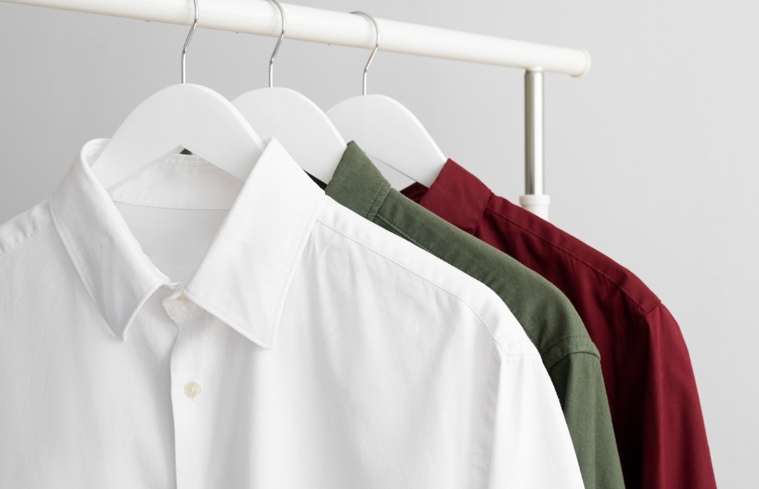 Three button-up shirts—one white, one green, and one burgundy—hang neatly on white hangers from a white clothing rack against a light background.