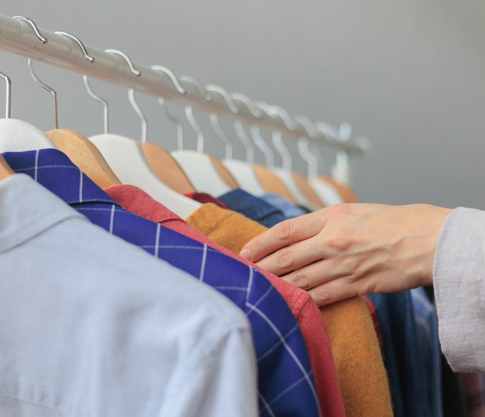 A person’s hand selects clothes from a row of shirts and jackets hanging on wooden hangers on a rack.