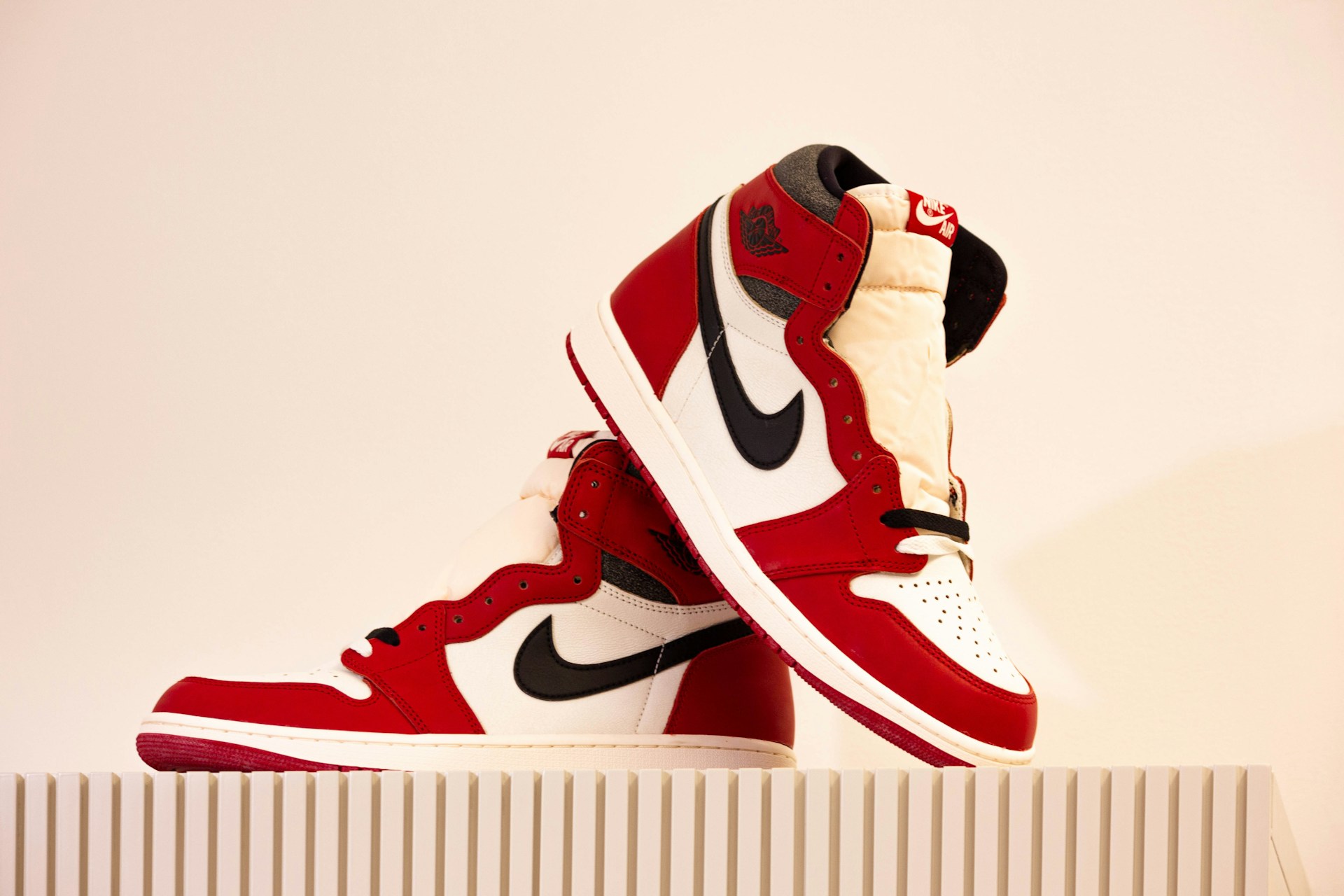 A pair of red, white, and black Nike Air Jordan 1 sneakers are displayed with one shoe propped on the other against a neutral background.
