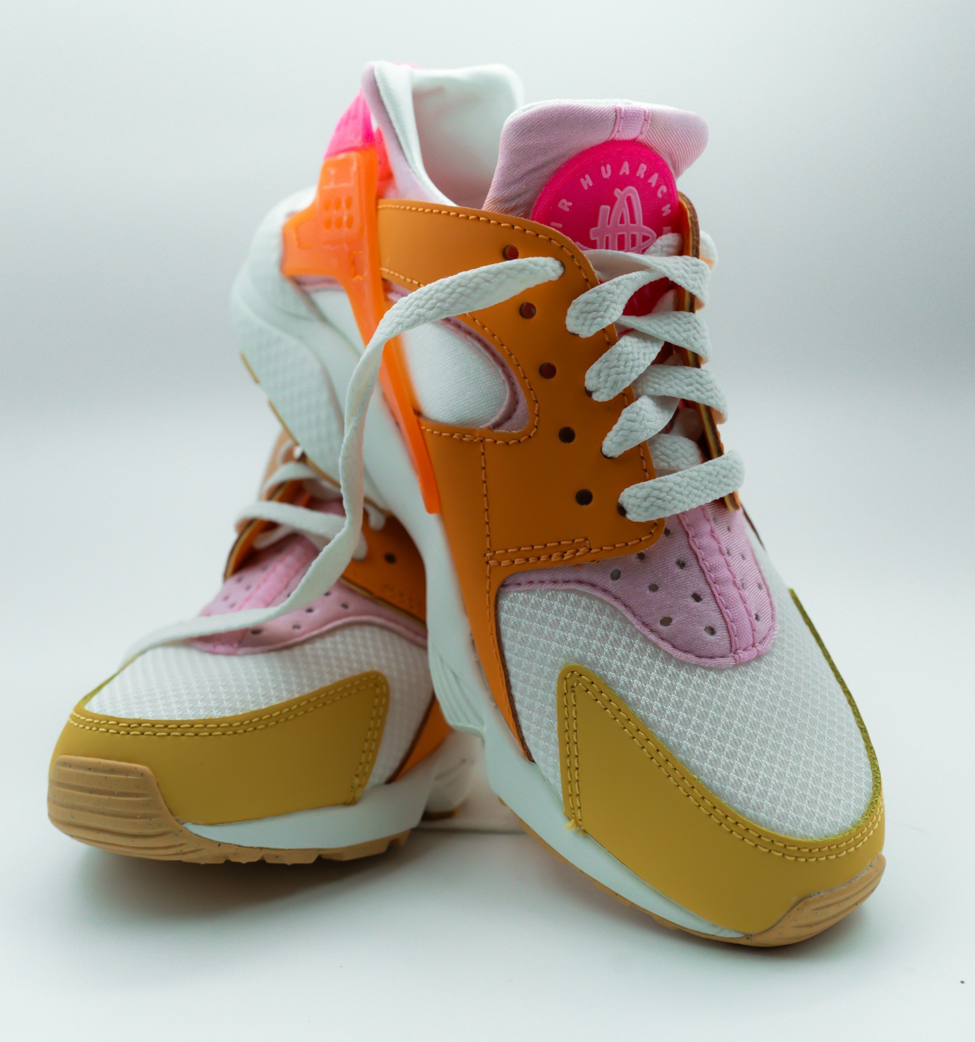 A pair of athletic sneakers featuring white mesh, yellow and orange accents, pink panels, and white laces, positioned with one shoe leaning against the other on a plain background.
