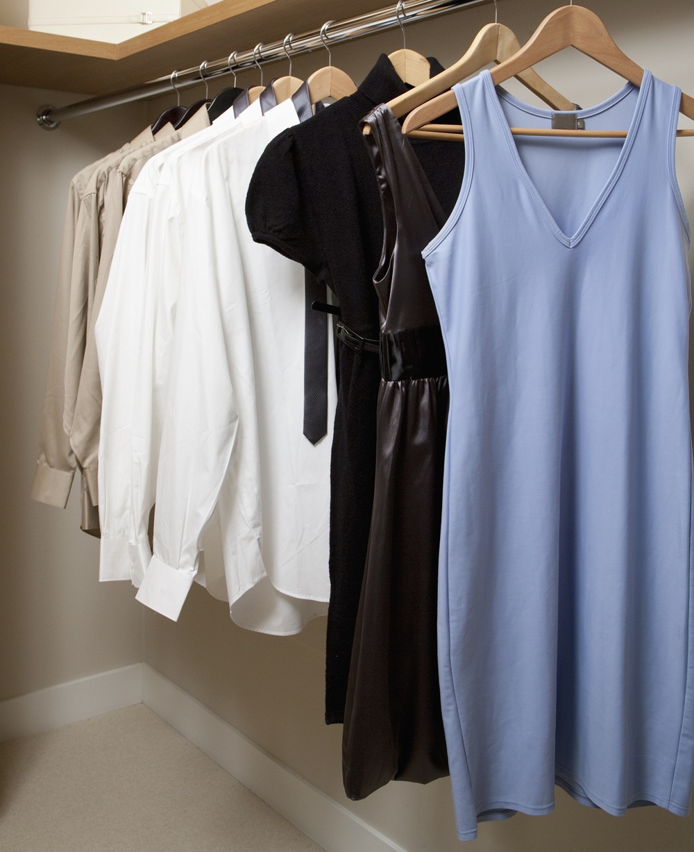 A row of assorted dresses, shirts, and a tie hanging on wooden hangers inside a closet.