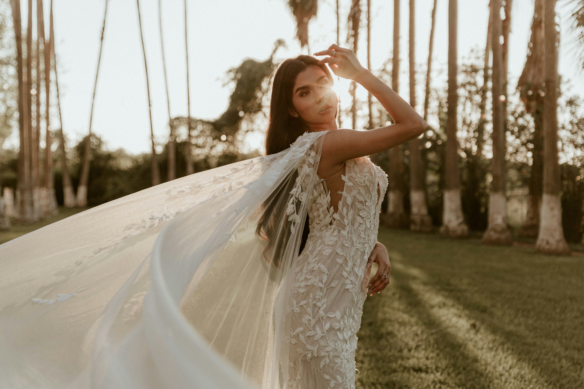 A woman in a detailed lace wedding dress stands outdoors among tall palm trees, with sunlight shining through her flowing veil.