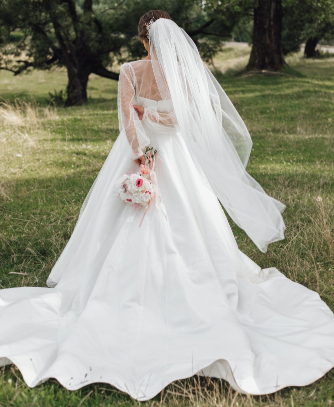 A bride stands outdoors on grass, facing away, wearing a long white wedding dress and veil, and holding a bouquet of flowers.