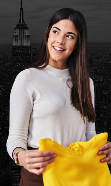 A woman in a white sweater holds a yellow garment and smiles, with a grayscale cityscape and an illuminated skyscraper in the background.