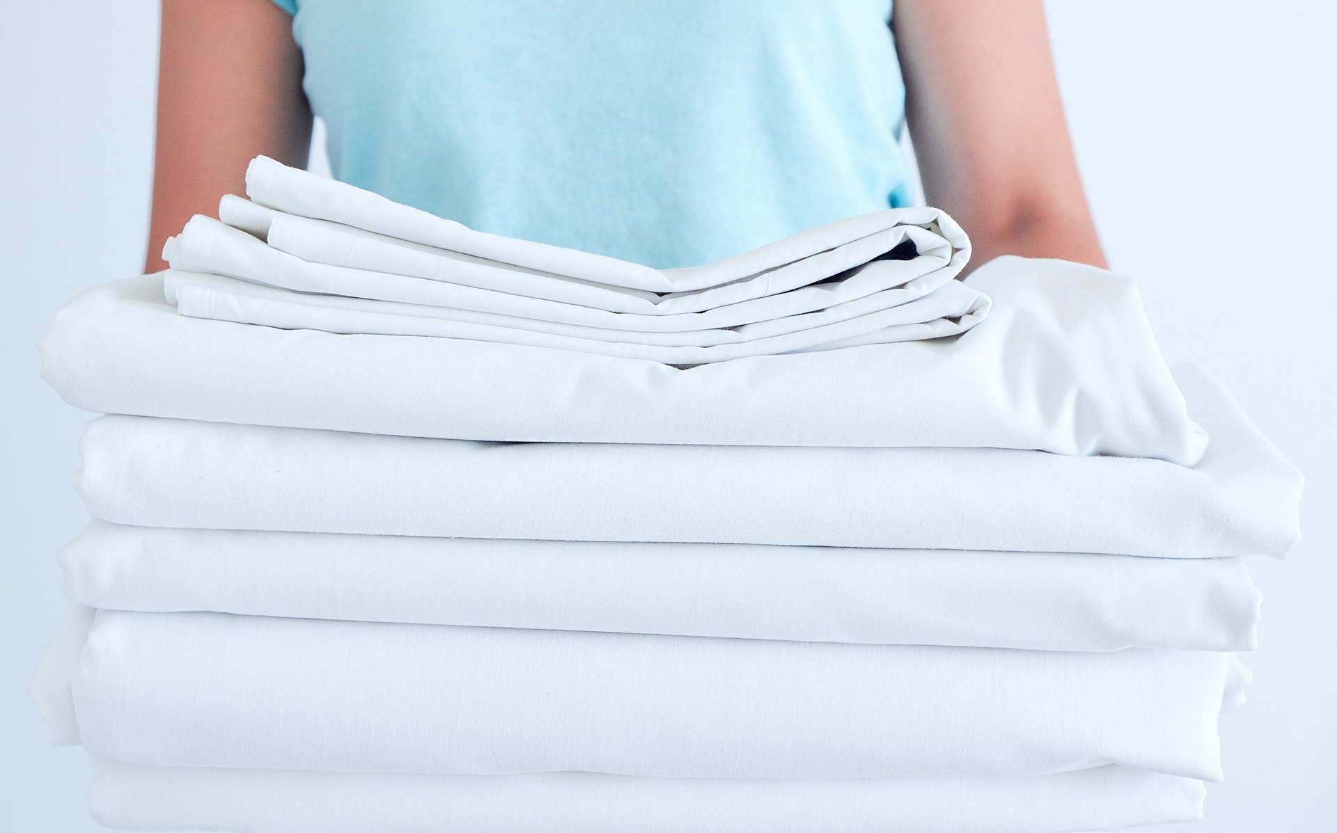 A person in a light blue shirt holds a stack of neatly folded white linens against a plain background.