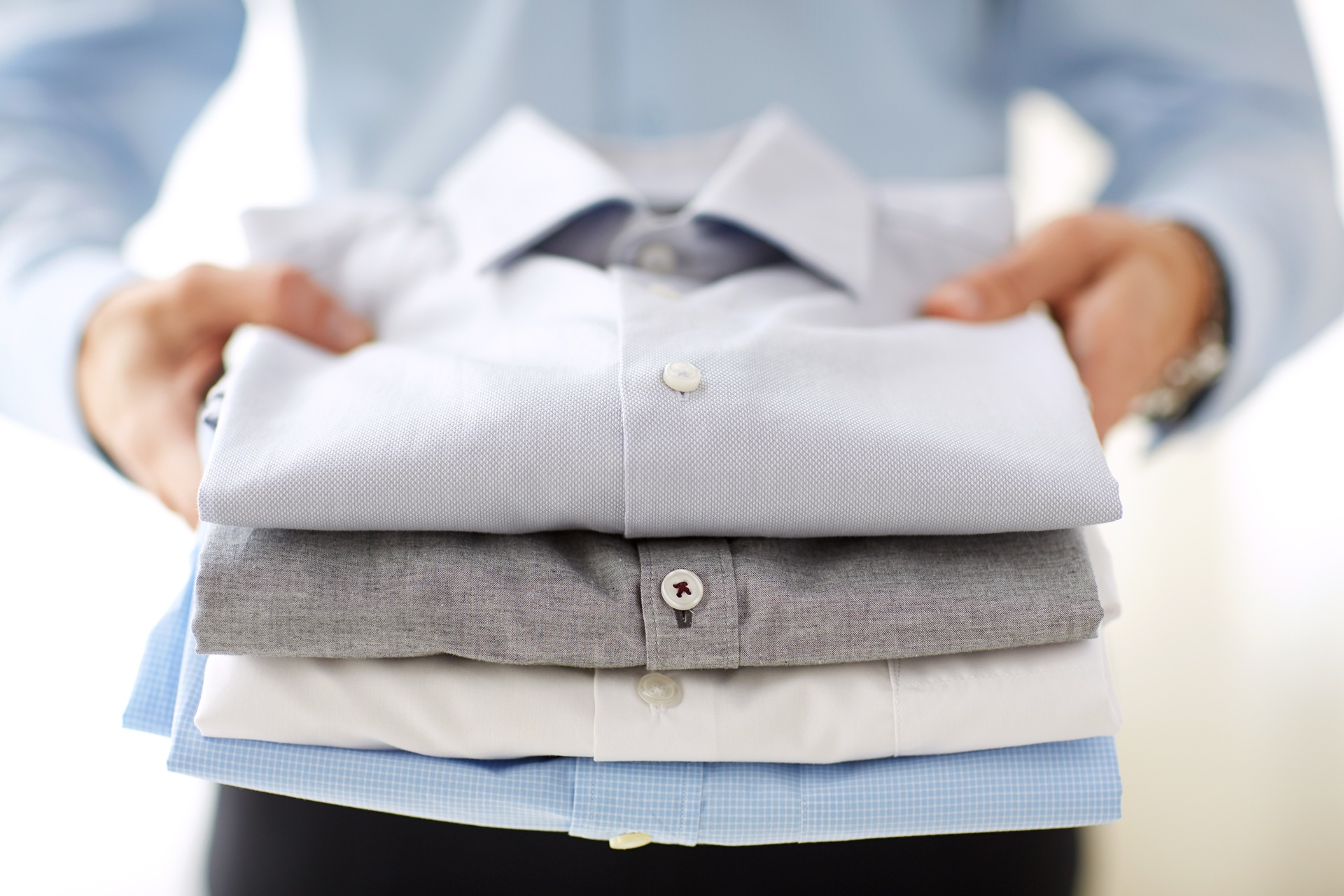 A person holding a neatly folded stack of four dress shirts in light colors, with the focus on the shirts and the person's hands.