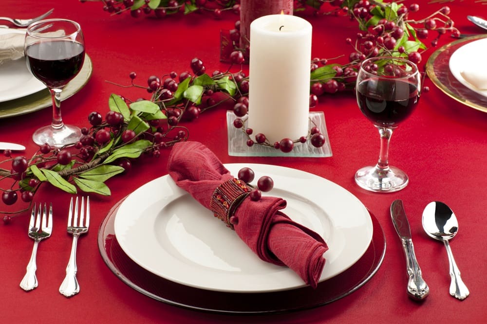 A formal table setting with a red napkin, white plate, silver cutlery, two glasses of red wine, a white candle, and red berry decorations on a red tablecloth.