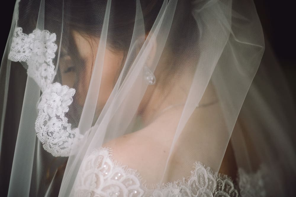 Close-up of a bride wearing a lace wedding dress and veil, with her face partly obscured by the veil and soft lighting highlighting the fabric details.