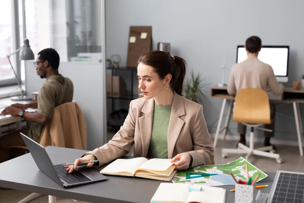 Woman in a beige blazer works at a desk with a laptop and open notebooks, while two other people work at desks in the background in a modern office setting.