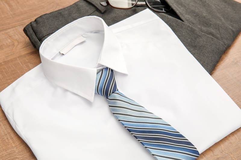 A neatly folded white dress shirt with a blue striped tie, gray trousers, and a pair of eyeglasses laid out on a wooden surface.